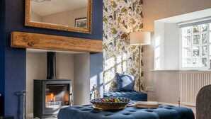Una sala de estar con chimenea, un espejo grande, papel tapiz estampado y un reposapiés azul con un frutero.
