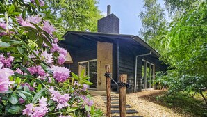 Un chalet rustique doté d’une cheminée, entouré d’une végétation luxuriante et de plantes en fleurs.
