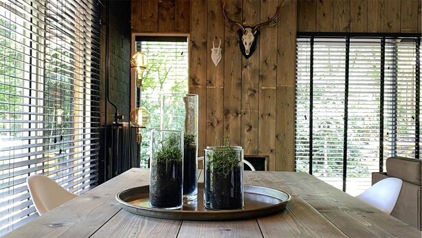 Une table à manger en bois, sur laquelle repose un plateau contenant deux vases en verre avec des plantes, le tout placé devant un mur en bois et agrémenté d’un crâne de cerf accroché au mur.