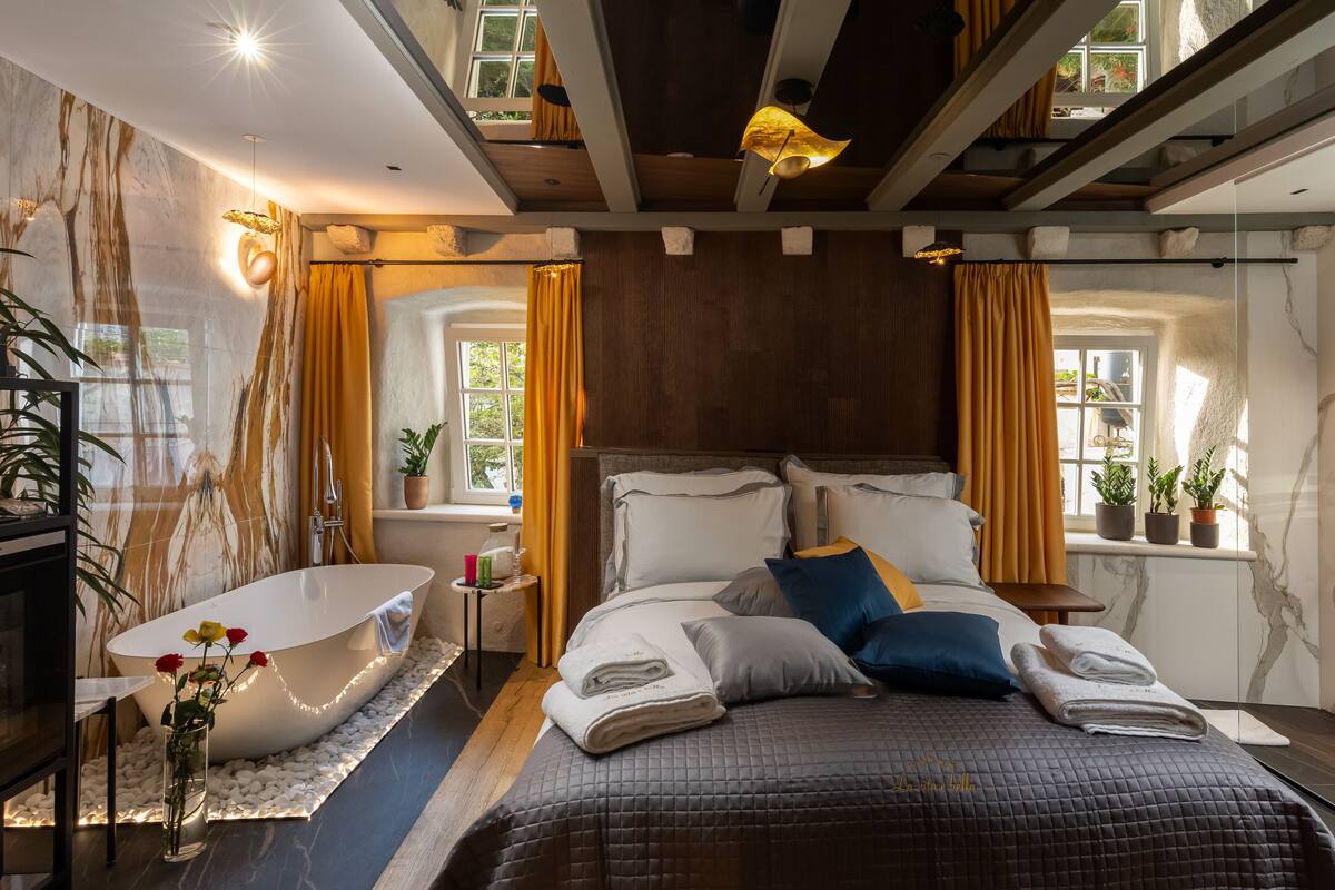 Un dormitorio moderno con una bañera grande, una cama con muchos almohadones y vistas al exterior a través de una ventana.