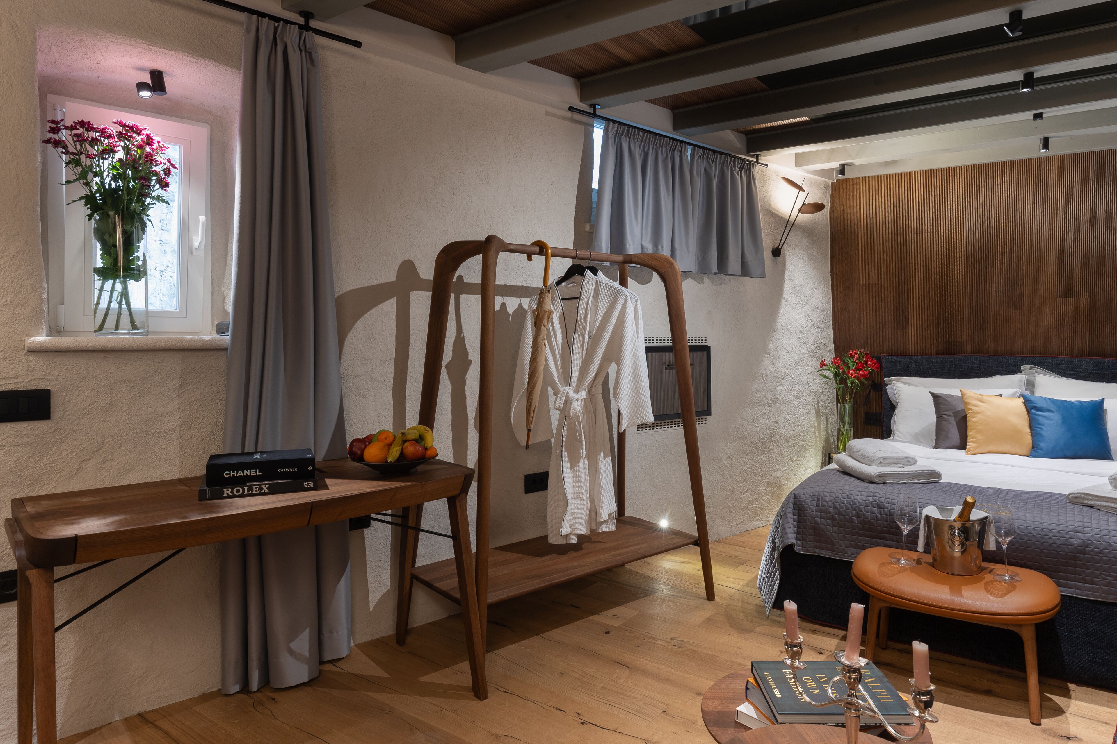 A bedroom with a wooden bed, a bedside table with a book and a bottle, a wooden chair, and a window with curtains.