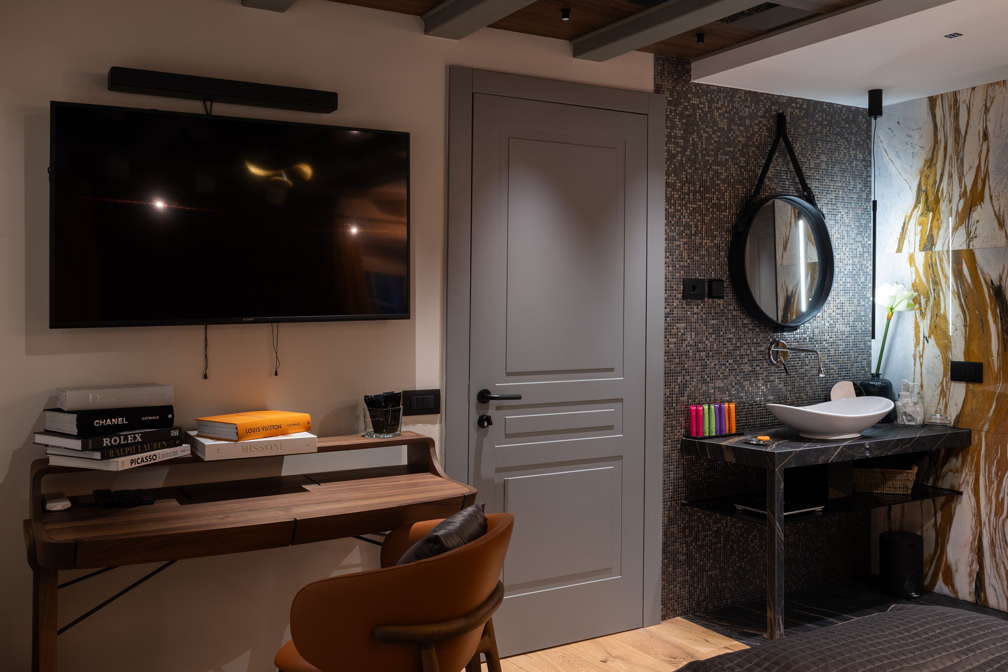 A modern room with a flat-screen TV, a wooden desk with books, a chair, a bathroom with a sink, mirror, and decorative tiles.