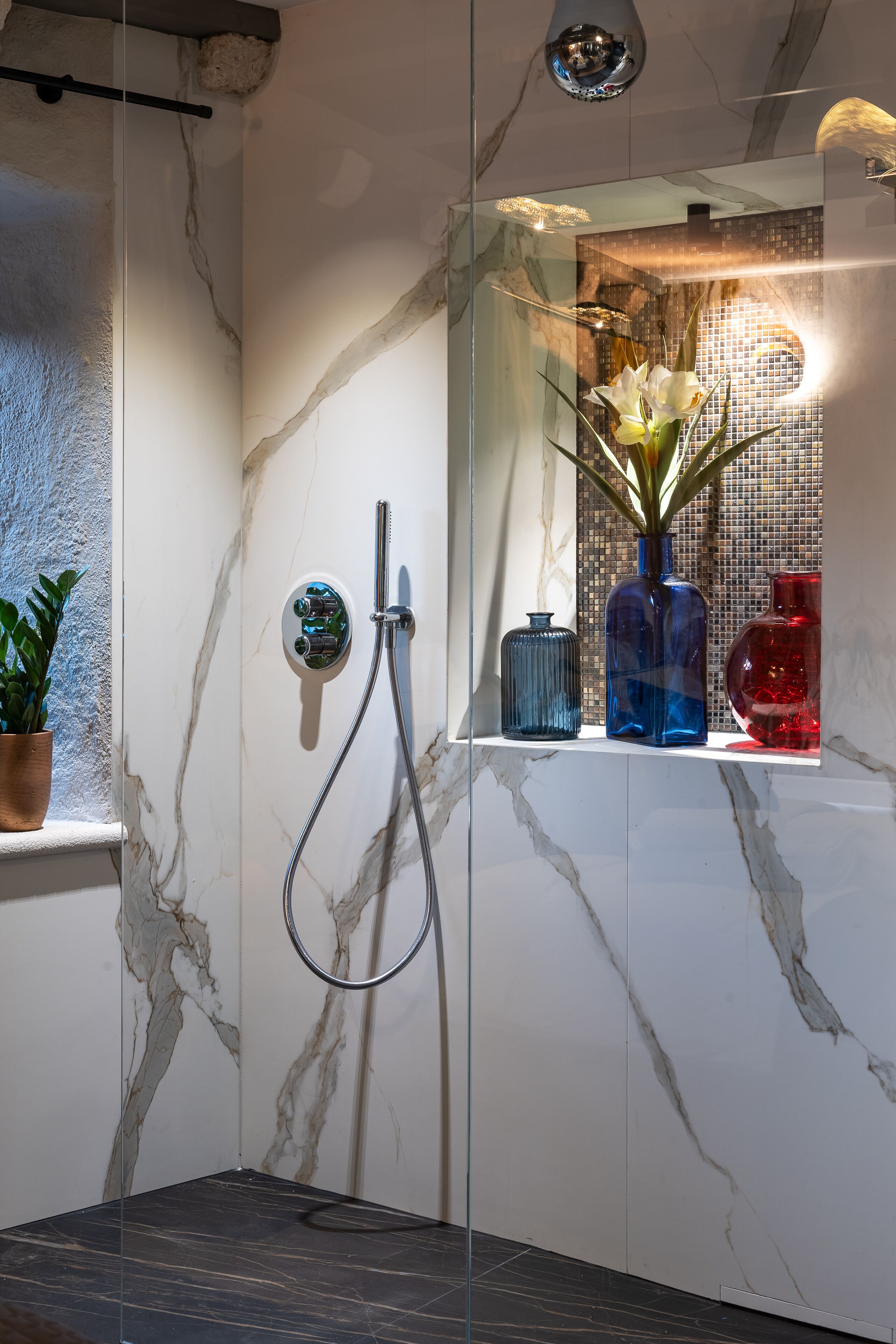 Un baño moderno con cabina de ducha de vidrio, paredes de mármol y un estante con jarrones decorativos.