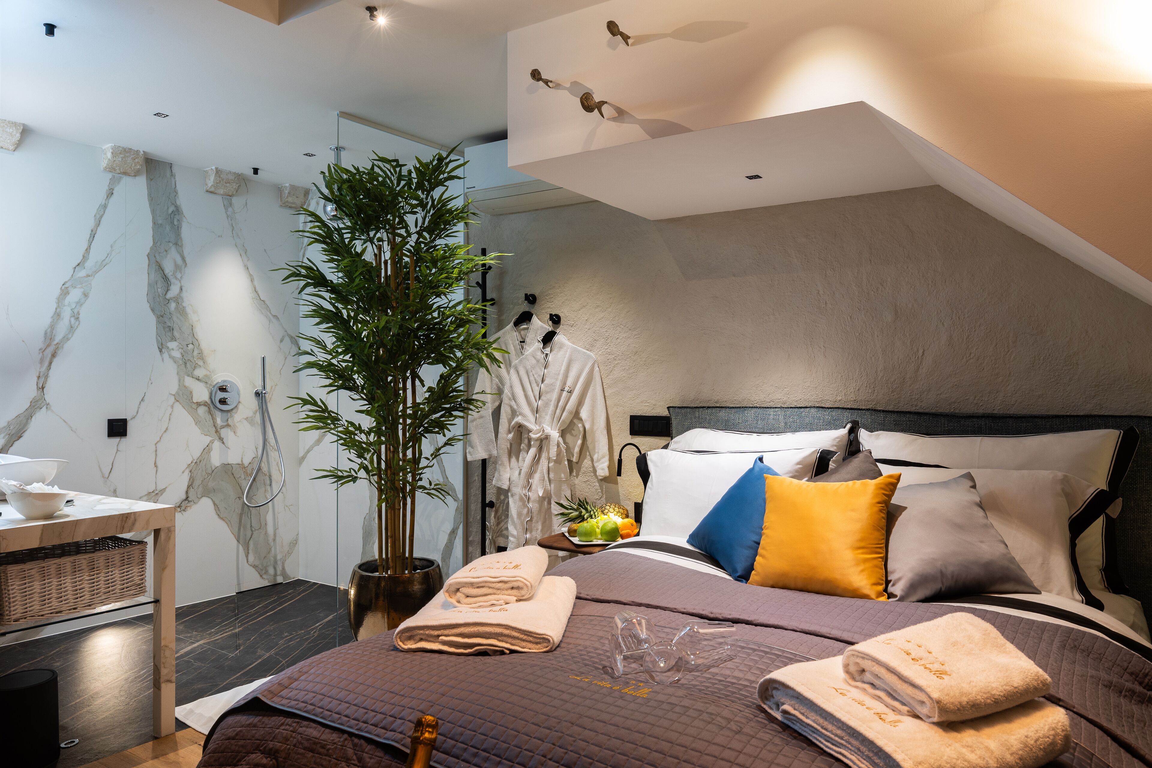A modern bedroom with a marble wall, a large bed with multiple pillows, a bedside table with a plant, and a nightstand.