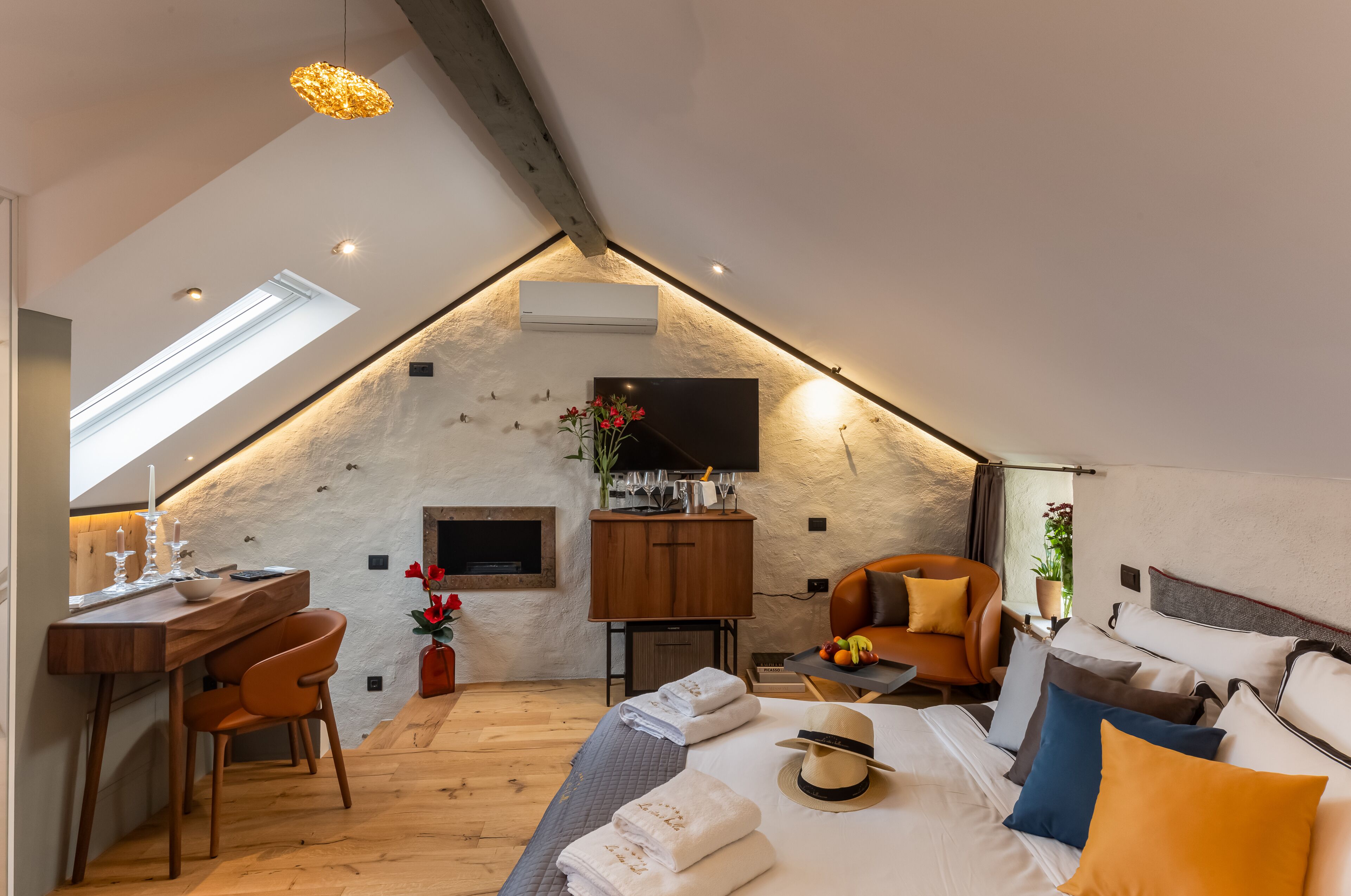 A cozy, modern loft bedroom with a sloped ceiling, a wooden desk, a comfortable chair, a bed with pillows, and a TV mounted on the wall.