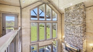 A spacious room with large windows, a stone fireplace, and a view of a green landscape. - Luxury 4 BR Cabin overlooking valley w/ Hot Tub, Game Room, Outdoor Fireplace (Sevierville)