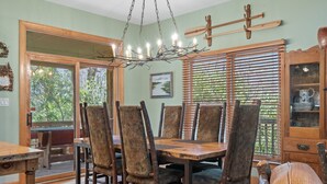 A dining area with a wooden table and chairs, a chandelier, and a view of greenery through the window. - The Lodge That Ruins Every Other Rental For You | Laughing Bear Lodge (Highlands)