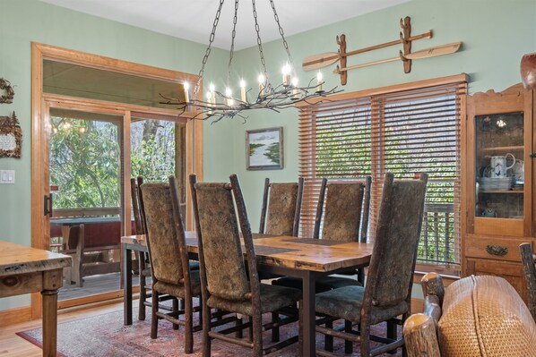 A dining area with a wooden table and chairs, a chandelier, and a view of greenery through the window.