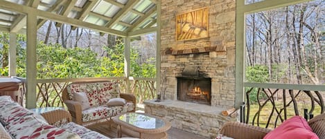 A covered outdoor seating area with wicker furniture, a stone fireplace, and a view of the surrounding trees.