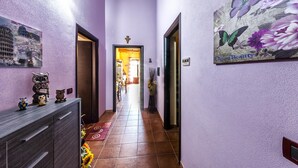 A narrow hallway with purple walls, a wooden ceiling, and a tiled floor. There are decorative owls on a cabinet, a painting of flowers, and a cross on the wall. - Affitacamere Domo - Luci (Nurachi)