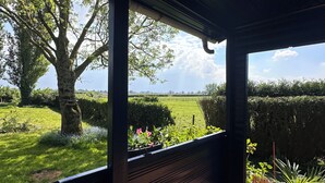 Ein überdachter Außenbereich mit einer Holzbank und Blick auf ein Wiesenfeld und Bäume.