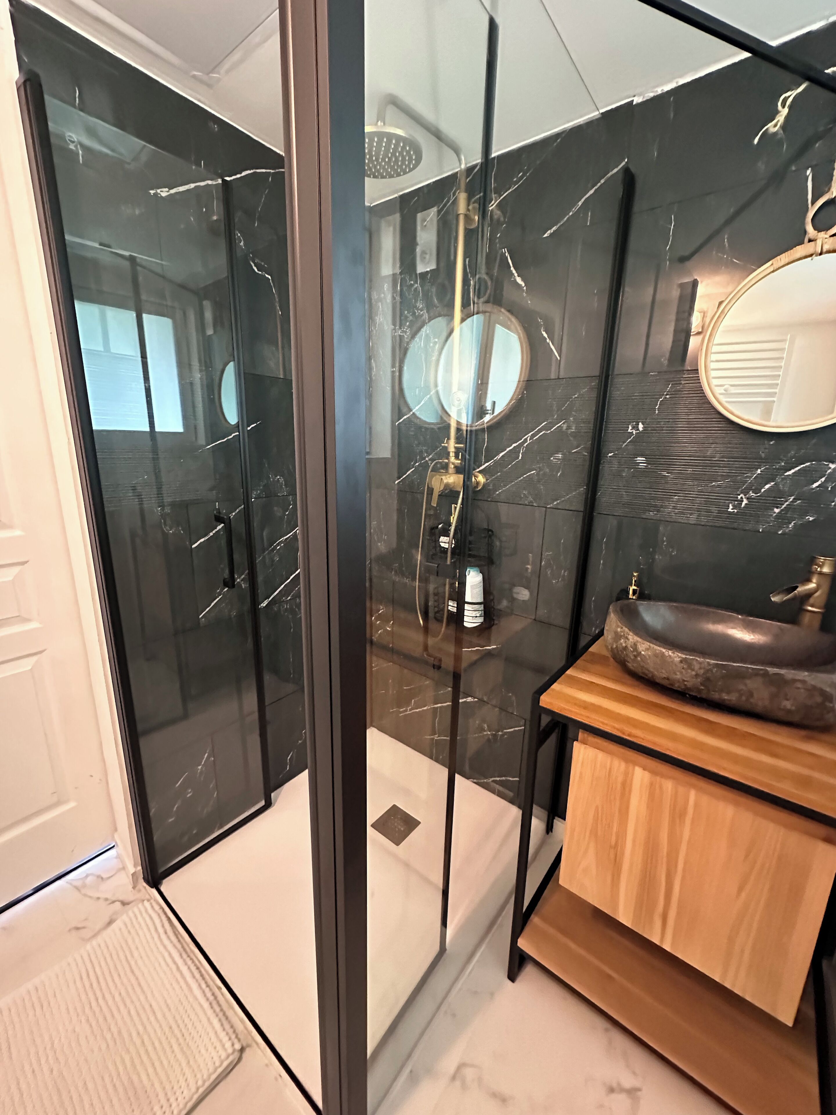 Une salle de bain moderne dotée d’une douche à parois de verre, d’un miroir rond et d’un meuble-lavabo en bois avec un vasque en pierre.
