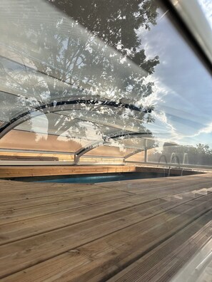 A pool area with a wooden deck, a covered seating area, and a view of trees and a clear sky. - Le Chalet de Margareth (Eecke)