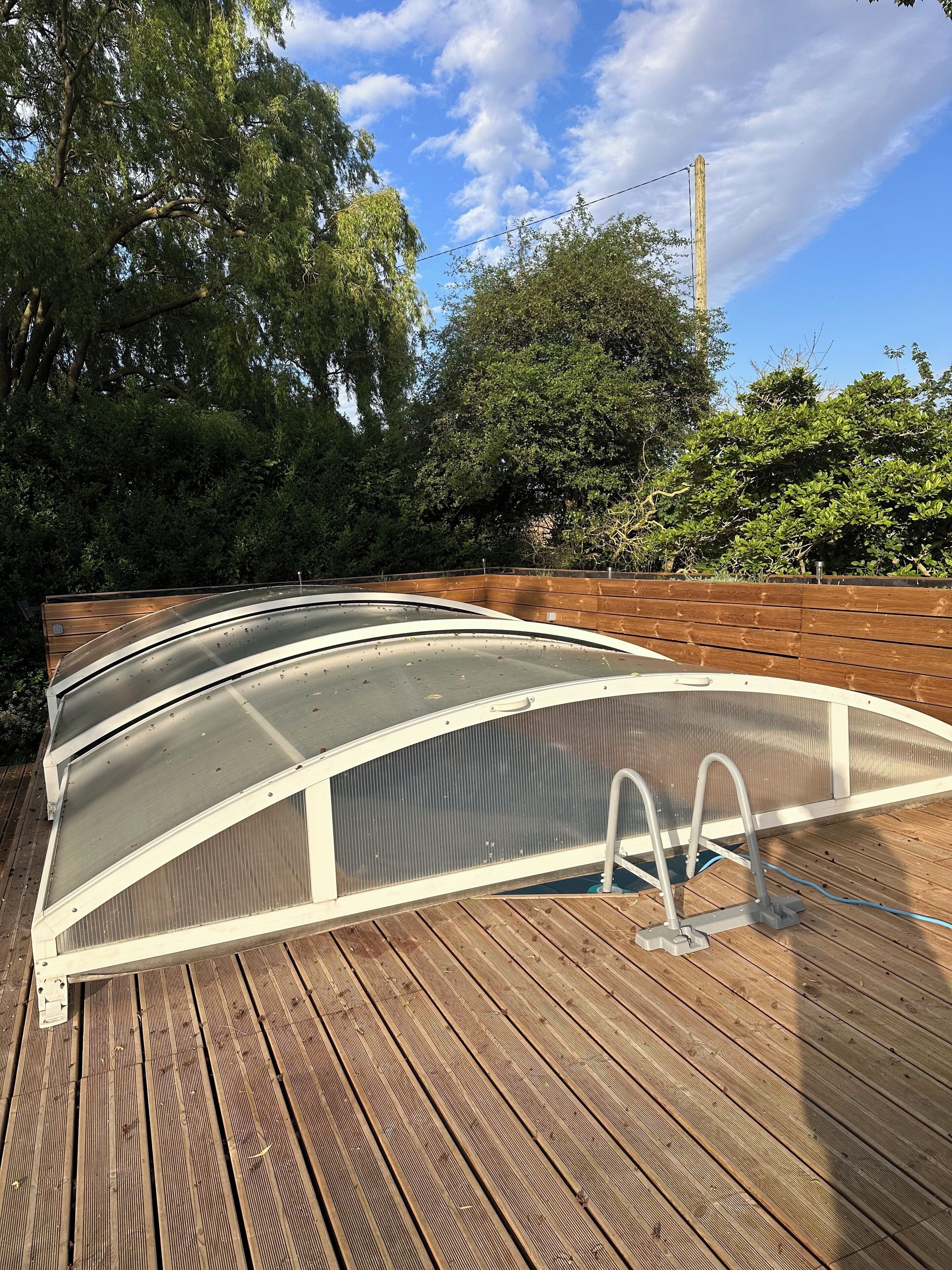 Una terrazza in legno con area piscina, circondata da alberi e un cielo sereno.