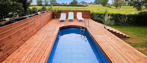 Una terrassa de fusta amb una piscina, tres gandules amb ratlles i vista a un paisatge verd.