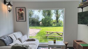 Ein modernes Wohnzimmer mit einem grauen Sofa, einem kleinen Tisch, einem Korbstuhl und einem karierten Teppich. Ein großes Fenster bietet Blick auf eine Wiese und Bäume.