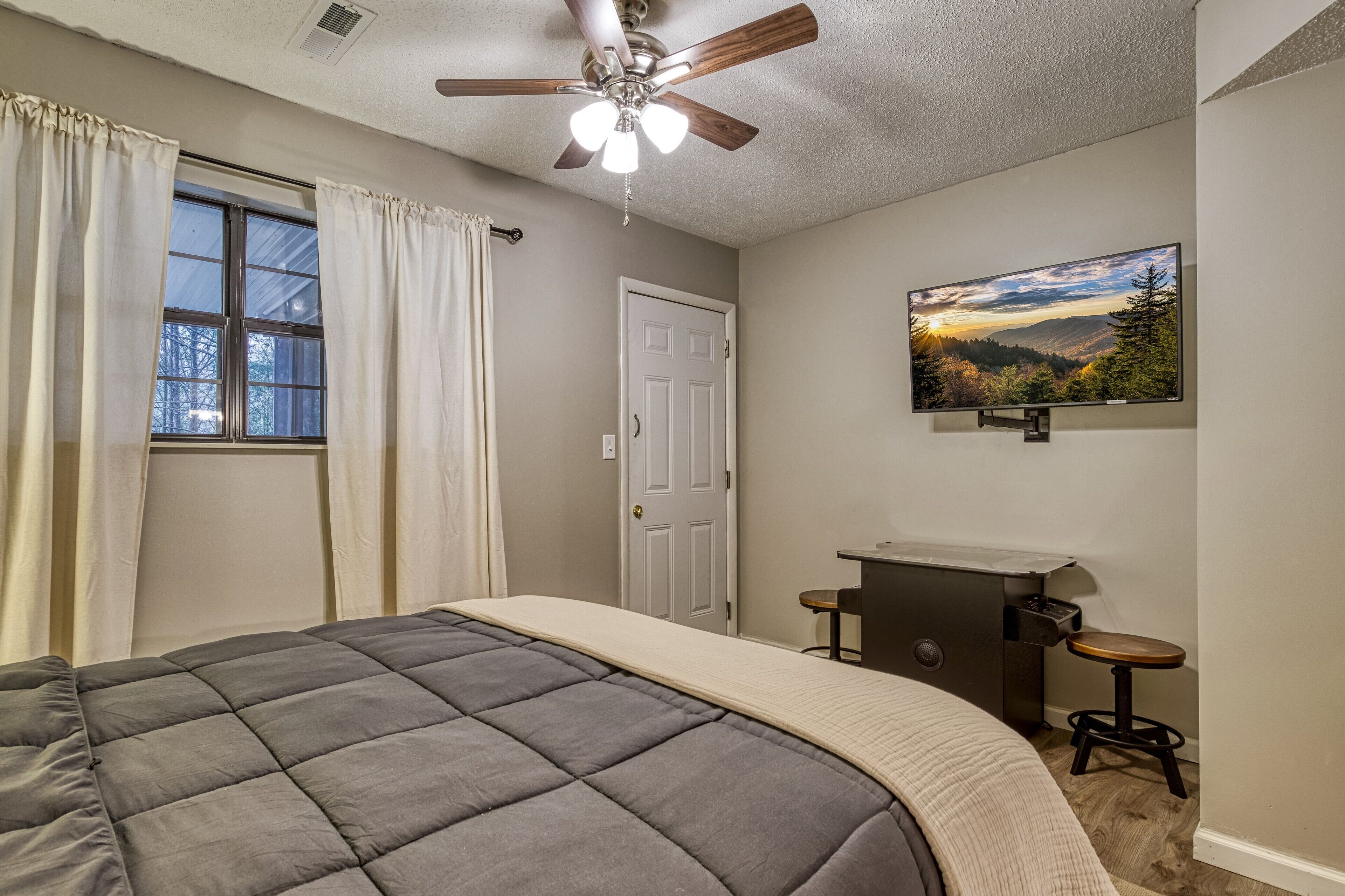 Un dormitorio con cama, ventilador de techo, ventana con cortinas, puerta, escritorio con silla y un cuadro en la pared.