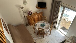 Una habitación pequeña con cama, una mesita de comedor con sillas, un televisor en un estante y vistas al exterior por una puerta corredera.