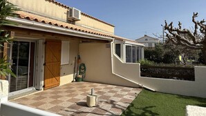 Une maison avec un patio carrelé, une porte en bois et une fenêtre donnant sur le jardin.