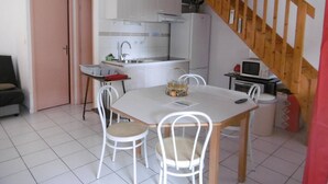 Une cuisine compacte avec des meubles blancs, un évier et un four à micro-ondes. On y trouve une table à manger avec des chaises et une petite table sur laquelle est posé un panier de fruits. Un escalier avec des rampes en bois est visible à l’arrière-plan.