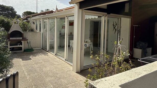 Un patio cubierto con puerta de vidrio que da a un espacio interior, una parrilla de barbacoa y un pequeño jardín.
