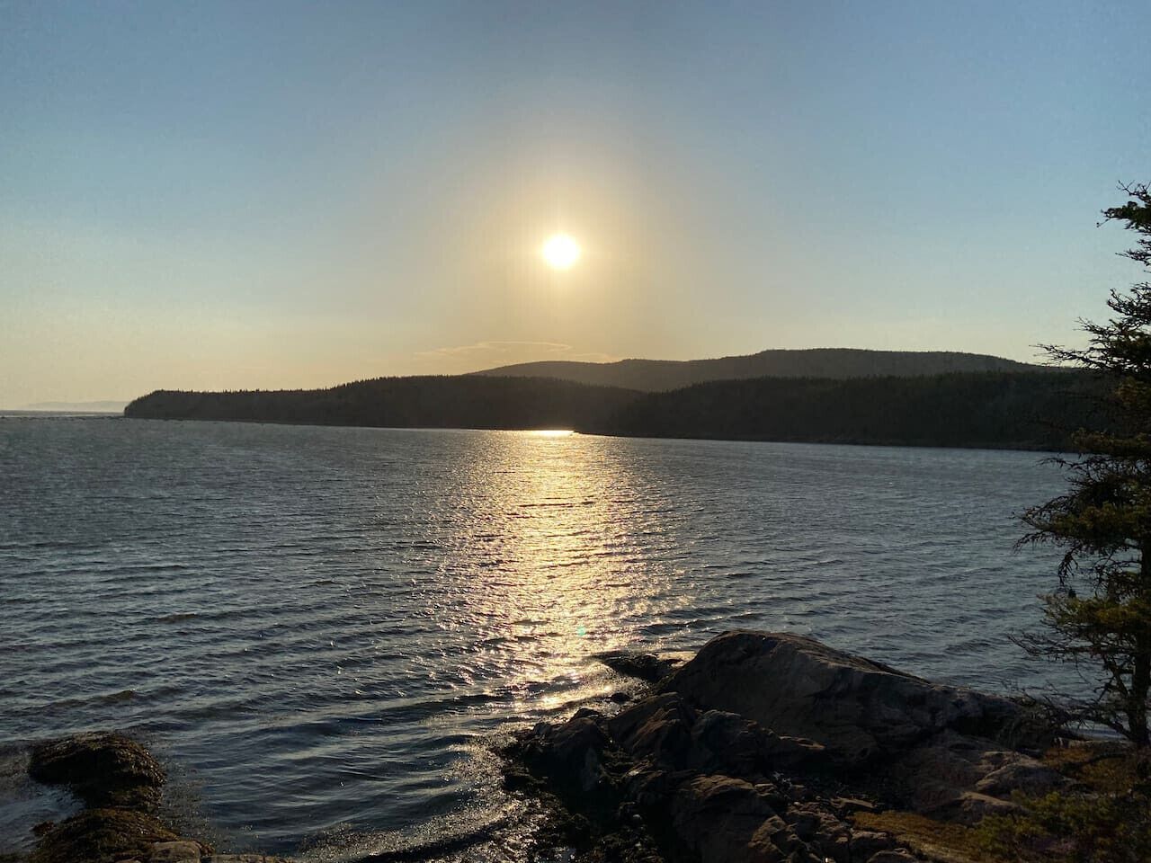 Un lac paisible, dont la surface reflète la lumière du soleil, bordé de rives rocheuses et entouré de collines lointaines.