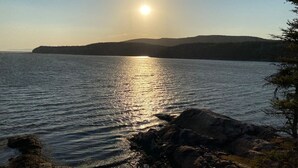 A calm lake with a sun reflecting on the water, surrounded by rocky shores and distant hills. - Le Havre du Pilote Aux Bergeronnes (Grandes-Bergeronnes)