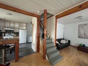 A kitchen with modern appliances, a dining area with a wooden table, and a staircase leading to a living area with a black sofa. - Le Havre du Pilote Aux Bergeronnes (Grandes-Bergeronnes)