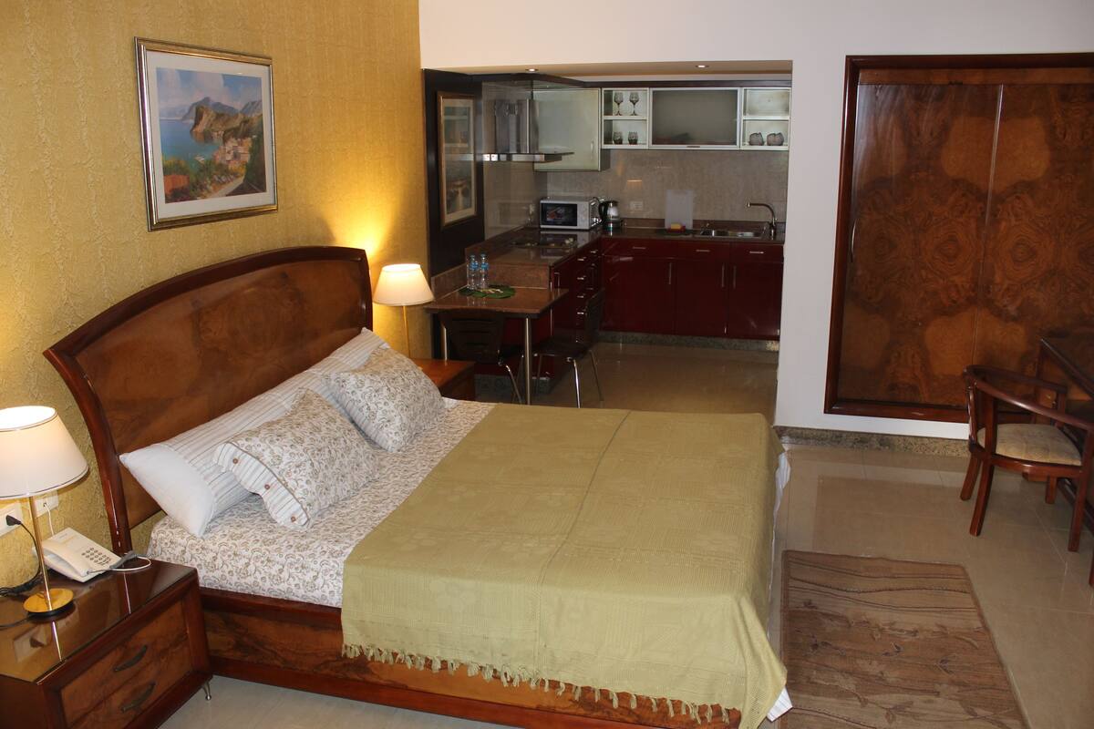 A hotel room with a bed, bedside table, lamp, and a view of the kitchen.