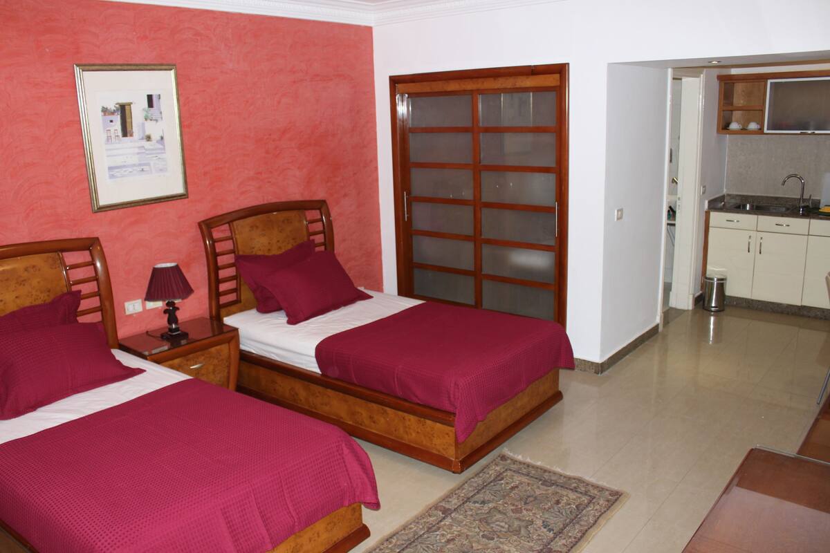 A hotel room with two wooden beds, a small bedside table with a lamp, a framed picture on the wall, and a view into the adjacent kitchen area.
