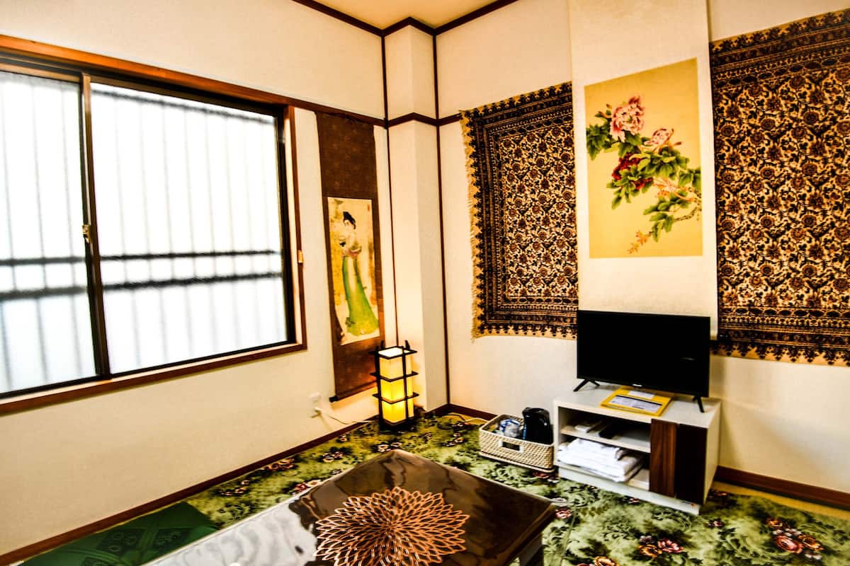 A traditional Japanese room with tatami flooring, a tatami mat in the center, a television on a wooden stand, and a window with a view of the outside.