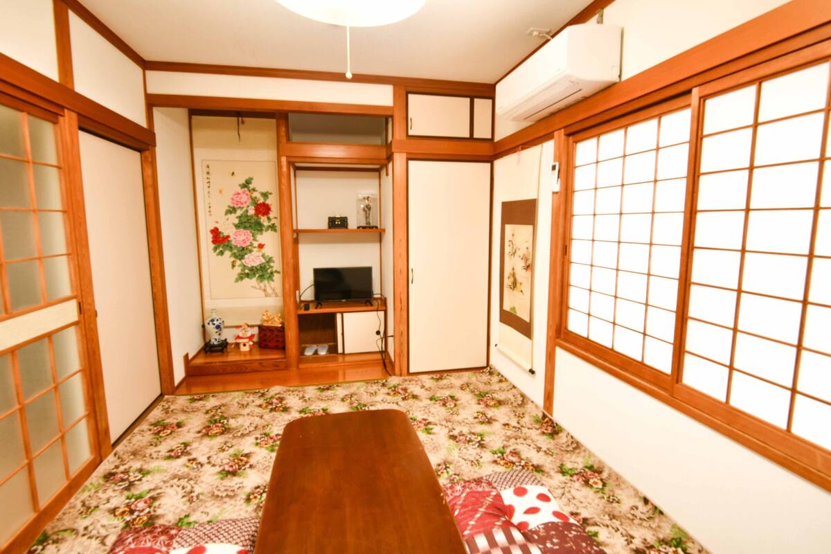 A traditional Japanese room with tatami flooring, a low wooden table, sliding doors, and a television on a shelf.