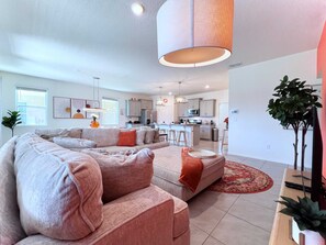 A modern living room with a sectional sofa, a kitchen in the background, and a large window.