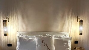 En pænt redt seng med hvide sengetøj og puder, herunder én med broderede initialer. To vægmonterede lamper giver blødt lys.