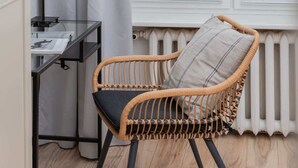 Une chaise en osier avec un coussin, une table en verre sur laquelle se trouvent des papiers, et un radiateur à l’arrière-plan.