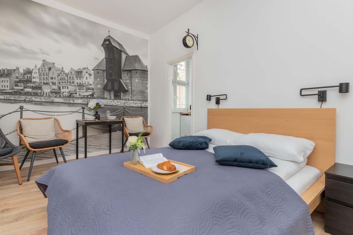Ein Schlafzimmer mit Bett, Holz-Kopfteil, Nachttisch, einer Wanduhr, einem Stadtbild in einem Bilderrahmen, einem Esstisch mit Stühlen und einer Ablage mit einem Croissant.