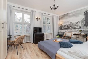 A modern hotel room with a large bed, a desk with a chair, a television, and a view of a cityscape through the windows. - Modern Studio in the Heart of Gdansk | A Place to Work (Gdańsk)