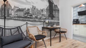 Une pièce chaleureuse décorée d’une fresque représentant un paysage urbain, avec un canapé en osier, deux fauteuils, une petite table surmontée d’un vase, et un coin cuisine doté de meubles blancs et d’un luminaire suspendu.