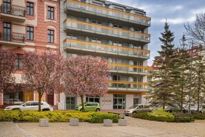 Un edificio residencial de varios pisos con balcones, un estacionamiento y un parque pequeño con árboles florecientes.