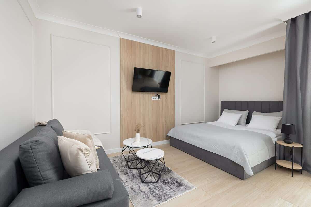 Une chambre d’hôtel moderne avec un canapé gris, un lit avec une literie grise, une petite table ronde avec un plateau blanc et un téléviseur fixé au mur.