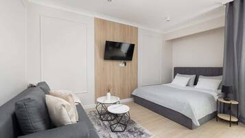 Une chambre d’hôtel moderne avec un canapé gris, un lit avec une literie grise, une petite table ronde avec un plateau blanc et un téléviseur fixé au mur.