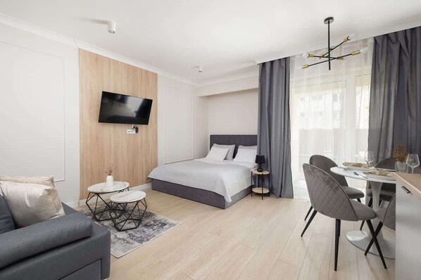 A modern hotel room with a bed, a TV, a dining area, and a large window with curtains.