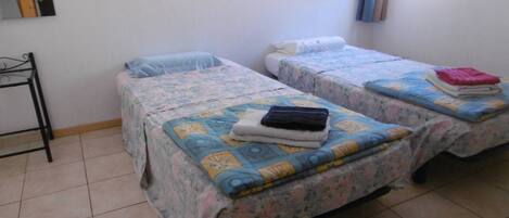 A room with two beds, each with a floral bedspread and a folded towel on top. There is a small table with a metal frame to the left of the beds.