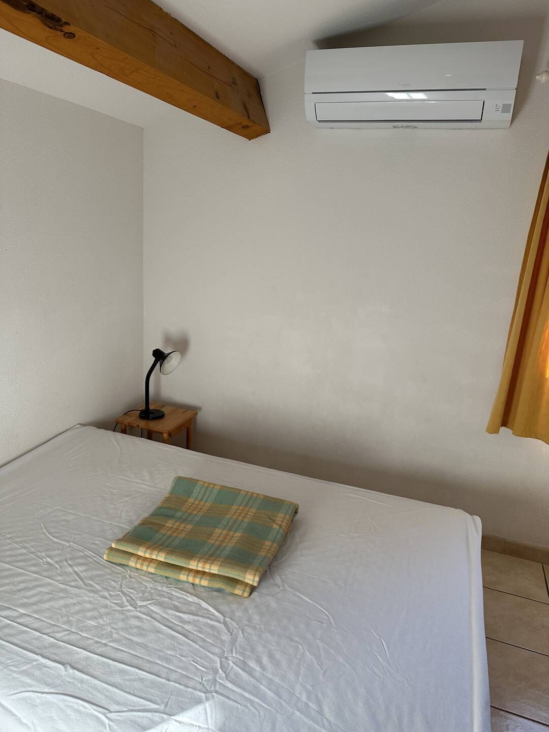Un dormitorio con cama, mesita de noche con lámpara, aire acondicionado y ventana con cortinas.