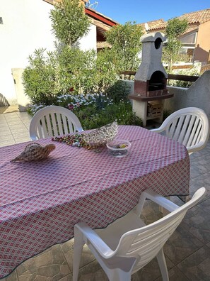 En udendørs patio med et bord dækket til to, omgivet af hvide stole, en stenovn og en have med forskellige planter og blomster.