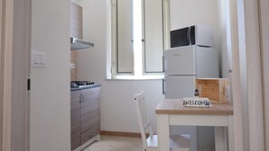 Une cuisine compacte équipée d’une cuisinière à gaz, d’un four à micro-ondes et d’un réfrigérateur. On y trouve une chaise en bois et une petite table sur laquelle est posée une pancarte indiquant « BIENVENUE ».