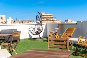 A rooftop terrace with wooden furniture, a hanging chair, and a view of city buildings. - Myia Apartment A by Wonderful Italy (Syracuse)