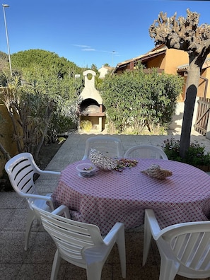 Une terrasse extérieure avec une table ronde et des chaises blanches, un four en pierre et un arbre dont l’écorce présente un motif particulier.