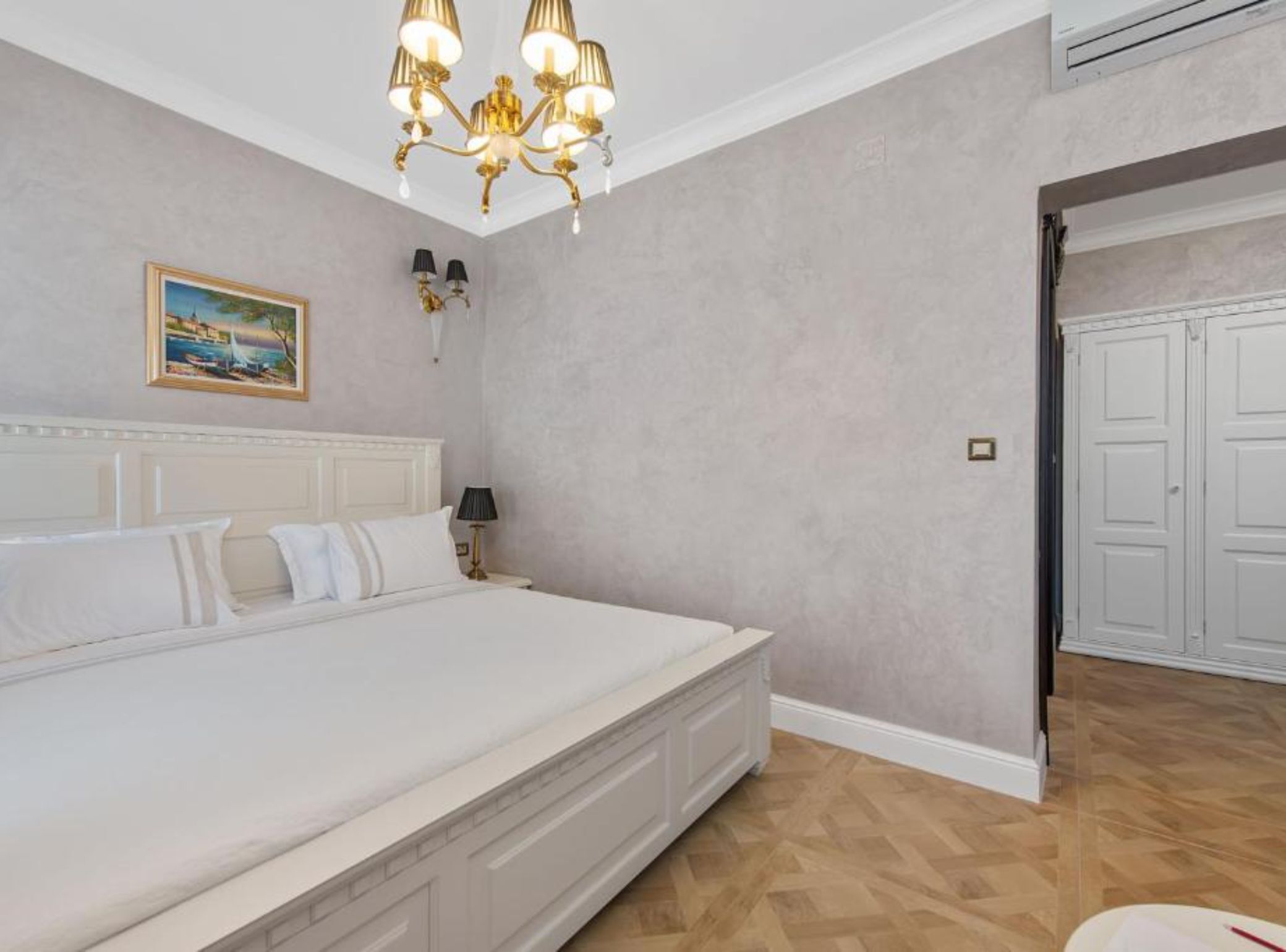 A bedroom with a large bed, a chandelier, a painting on the wall, and a door to the right.