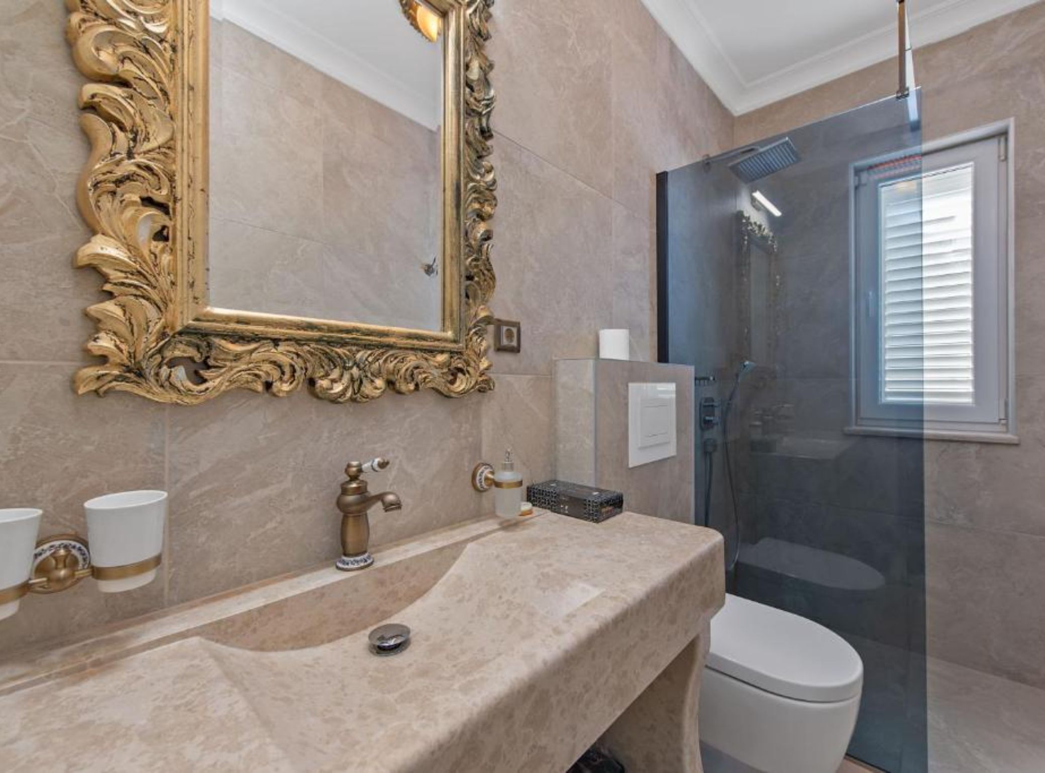 Un baño con lavamanos de mármol, un espejo ornamentado dorado, inodoro y ducha con mampara de vidrio.
